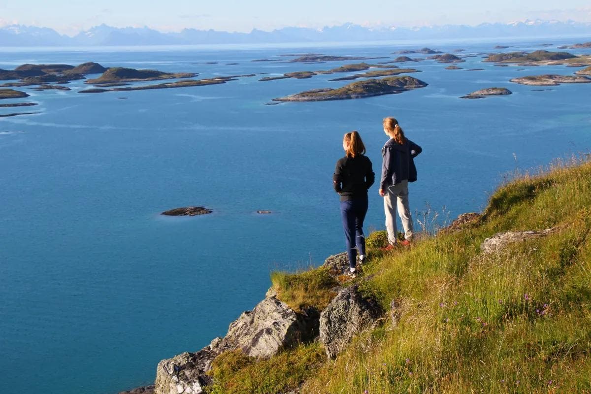 To personer står på en gresskledd fjellskrent og ser ut over et blått hav med mange små øyer og fjell i bakgrunnen.