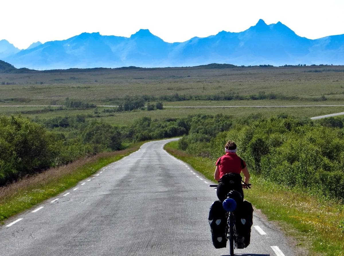En person på sykkel med store sidevesker sykler langs en asfaltert vei som strekker seg mot et flatt landskap med blå fjell i det fjerne.