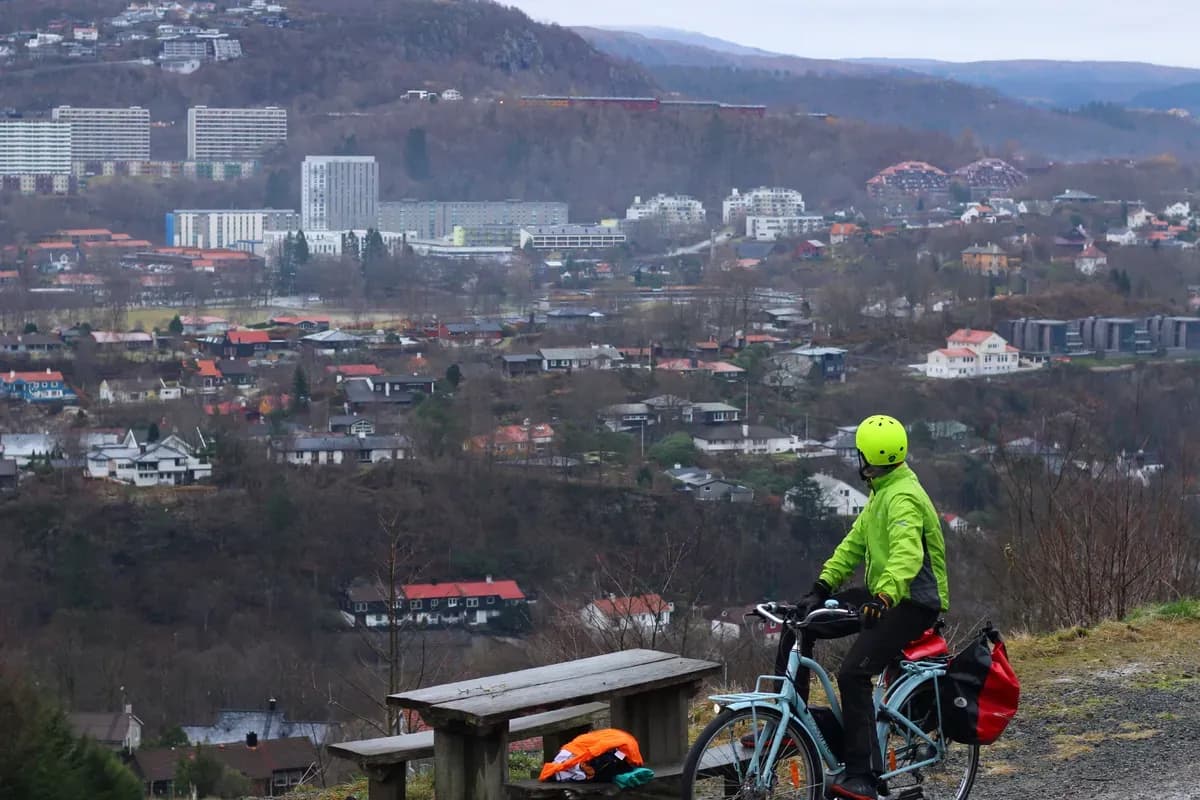En syklist med grønn jakke og gul hjelm ser utover et vidstrakt landskap med variert bebyggelse og skogkledde åser.